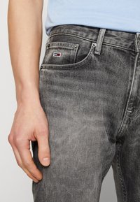 Jeans de mezclilla gris con ajuste slim, con un pequeño parche de logo en el bolsillo y una textura sutilmente desgastada. Mano posicionada en la cadera.