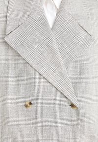 Dubbelzijdige blazer in een grijs-wit geruit patroon. Voorzien van grote revers en twee ronde, tan kleurige knopen aan de voorkant.