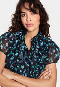 Črna prozorna bluzica z zelenim in modrim cvetličnim vzorcem, nabranim ovratnikom in rokavi, z gumbnim zapiranjem in lahkotno teksturo.