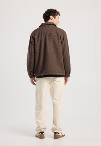 Brown wool jacket with a collared neck, side pockets, and loose fit, worn over a black top and cream-colored pants.
