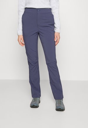Persoon draagt recht gesneden donkerblauwe broek, grijs topje met lange mouwen en grijze wandelschoenen, staand tegen een effen achtergrond.