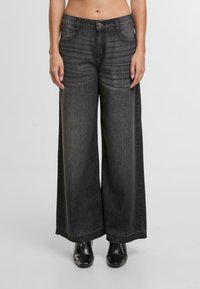 Černé denimové kalhoty se širokým střihem a vybledlým designem, předními kapsami a zapínáním na knoflík. Látka má hladkou texturu a rovný lem.