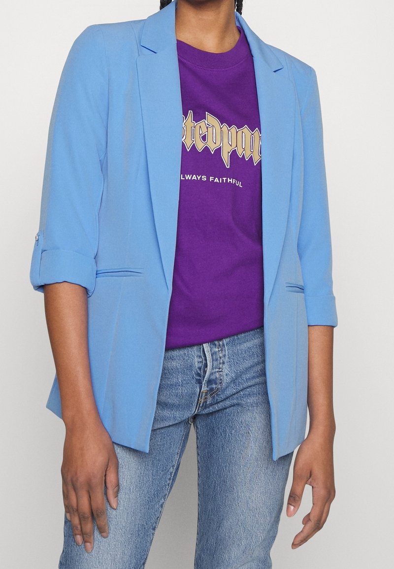 Personne portant un blazer bleu clair avec les manches retroussées sur un t-shirt violet à motif graphique et un jean bleu, sur un fond uni.