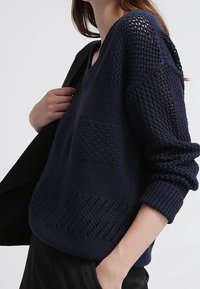 Námořnicky modrý pletený svetr s výstřihem do V, zdobený texturovanými vzory, žebrovanými manžetami a volným střihem; nošený s černou bundou.