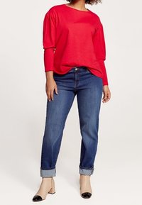 Punainen pitkähihaine paita, jossa on leveä pääntie, yhdistetty sinisiin käännettyihin farkkuihin ja beigeihin korkokenkiin, joissa on mustat kärkikappaleet. Sileä kangas.