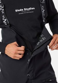 Schwarze Latzhose mit verstellbaren Trägern, ausgestattet mit einem Reißverschluss vorne und seitlichen Taschen. Texturiertes Material mit Logo-Akzenten und sichtbaren Pflegehinweisen.