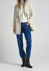 Beige double-breasted coat with large buttons, worn over a light sweater and blue jeans. Finished with black ankle boots.