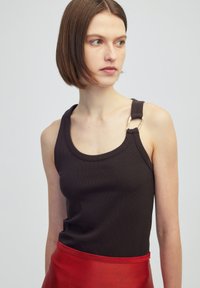 Zwarte geribbelde tanktop met brede banden, voorzien van een gouden metalen accent op één band. Model draagt een contrasterende rode rok.