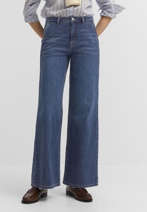 Person trägt hochtaillierte, weit geschnittene blaue Jeans, braune Loafers und ein hell gestreiftes Hemd mit den Händen in den Taschen vor weißem Hintergrund.