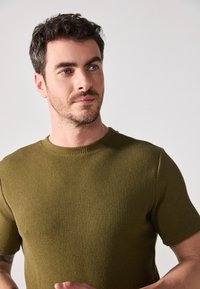 Camlă din tricot cu mâneci scurte, de culoare verde măsliniu, având un gât rotund și un material texturat. Design casual cu o siluetă ajustată.