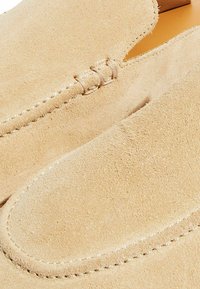 Scarpe slip-on in suede beige con punta rotonda, caratterizzate da cuciture decorative e una texture liscia. Senza loghi o decorazioni visibili.