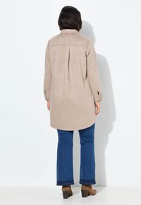 Beige Wildlederhemdjacke mit einer Rückensaumdetail, langen Ärmeln und seitlichen Schlitzen, kombiniert mit blauen Schlagjeans und braunen High-Heels-Stiefeln.