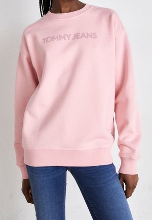 Osoba ubrana w jasnoróżową bluzę z logo "TOMMY JEANS" i niebieskie dżinsy, stojąca na tle jednolitego białego tła.
