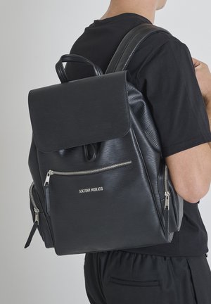 Mochila de piel negra con una superficie texturizada, tapa con solapa, bolsillo frontal con cremallera y branding. Cuenta con correas de hombro ajustables y un asa.
