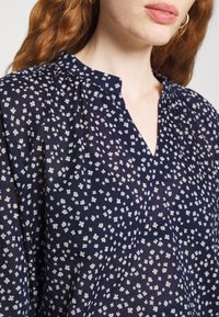 Blusa floreale blu navy in tessuto leggero, con scollatura a V e piccoli motivi floreali bianchi sparsi. Dettaglio in piqué sulla parte superiore.