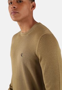 Maglione a maglia verde oliva con scollatura rotonda, polsini a costine e un piccolo logo ricamato sul lato sinistro del petto. Texture morbida e design aderente.
