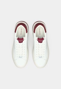 Hvite sneakers med en glatt skinnoverdel, røde semskede detaljer på tungen og hælen, rund tå og flate gummisåler. Merket med "GUESS."