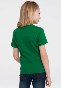 Green cotton T-shirt with short sleeves, round neckline, worn with dark denim jeans featuring pink patterned stitching on the pockets.