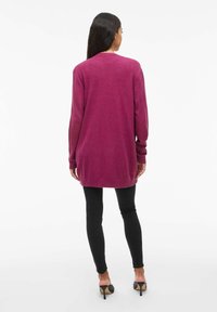 Długi magenta sweter z miękkiej dzianiny, z ribbowanymi mankietami i dołem, zestawiony z czarnymi legginsami i butami na obcasie.
