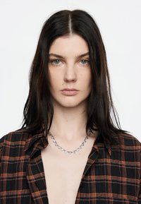 Collar de plata con cristales en forma de corazón, descansando sobre un cuello desnudo. El sujeto lleva una camisa de cuadros negra y naranja.