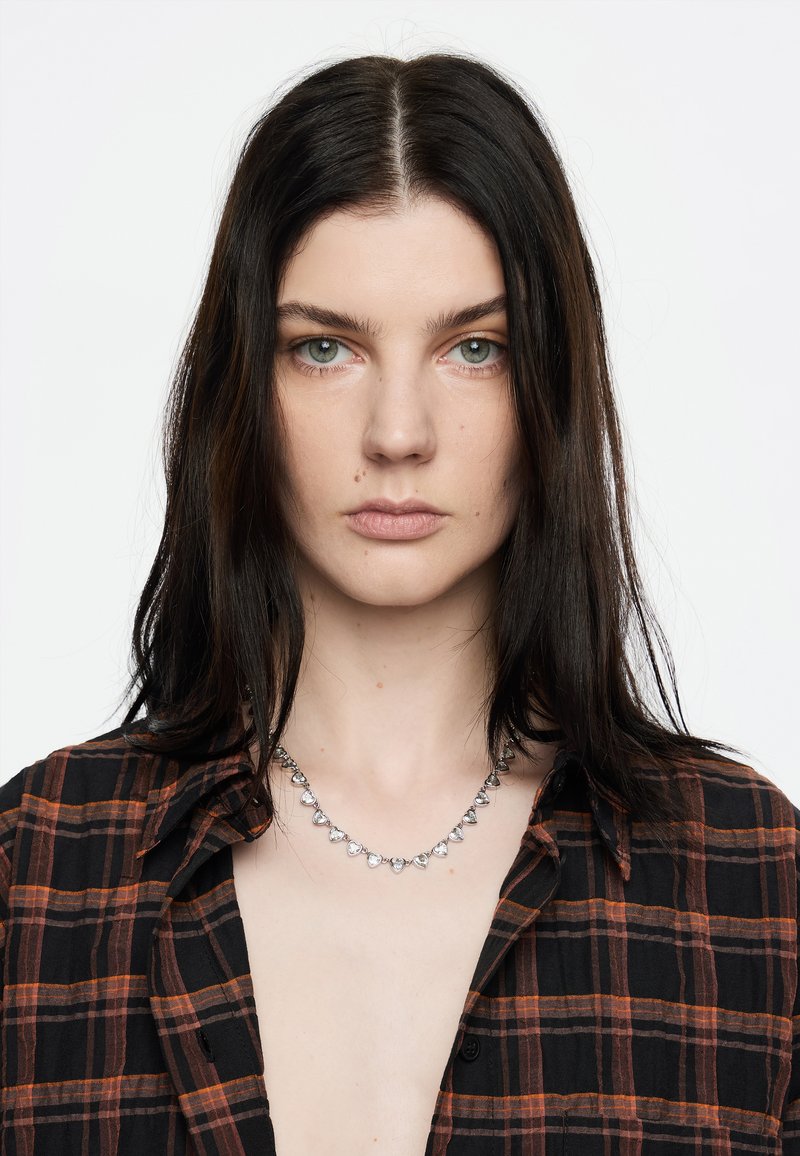 Collar de plata con cristales en forma de corazón, descansando sobre un cuello desnudo. El sujeto lleva una camisa de cuadros negra y naranja.