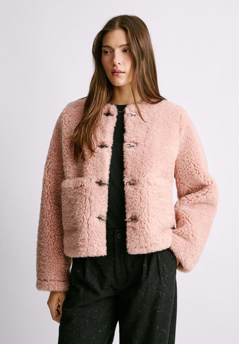 Giacca in pelliccia artificiale rosa con una forma quadrata, maniche lunghe, grandi tasche frontali e chiusure metalliche al centro. Indossata sopra una maglietta nera.