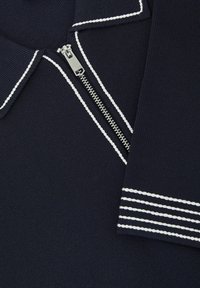 Marineblåt strikket stof med hvidt kantbånd, sølvlynlås halvt lukket, og hvidstribet manchet-detalje på ærmet.