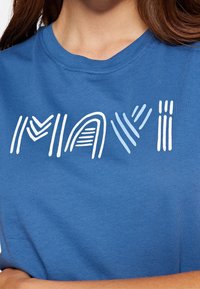 Modré bavlněné tričko s bílým stylizovaným nápisem "MAVI" s různou tloušťkou a tvary linií, kulatým výstřihem a krátkými rukávy.