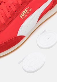 Red sneaker featuring a suede and nylon upper, white leather accents, gold logo, with two white flat shoelaces beside it.