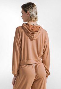 Kobieta o krótkich jasnych włosach ubrana w luźną brązową bluzę z kapturem i dopasowane spodnie, ukazująca tatuaże motyli na karku, stoi bokiem.