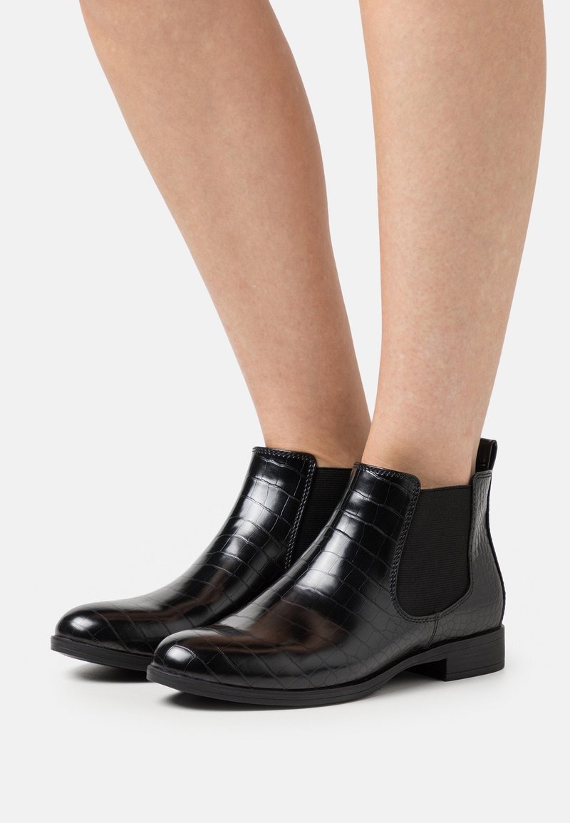 Bottines noires brillantes à motif crocodile avec panneaux élastiques sur les côtés, portées sur des jambes nues sur fond blanc.