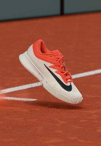 Παπούτσι τένις Nike με λευκό επάνω μέρος, πορτοκαλί λεπτομέρειες, μαύρο λογότυπο, υφή στην σόλα, επένδυση στη μεσαία σόλα και διασκορπισμένα σωματίδια πηλού.
