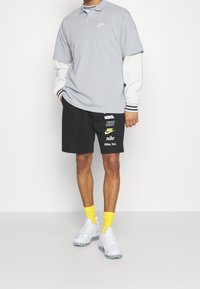 Muž oblečený v vrstvených šedobílých dlouhých rukávech a černých Nike šortkách s několika bílými a žlutými logy Nike, žlutými ponožkami a bílými teniskami.