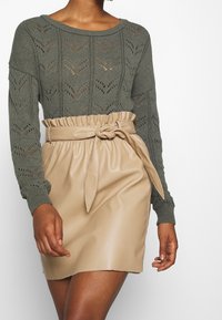 Pull en maille vert foncé avec motif en point ajouré ; jupe en similicuir beige avec ceinture nouée, texture lisse et design à couches.