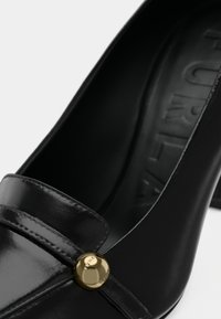 Pantof de piele neagră cu un finisaj neted, având un buton decorativ auriu și un toc mic. Design elegant, rotunjit, cu vârful închis.