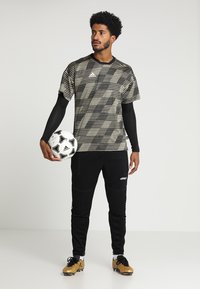 Zwart en beige gestreepte korte mouwen jersey, zwarte lange mouwen, zwarte gestructureerde joggers, met een voetbal in de handen, en gouden voetbalschoenen.