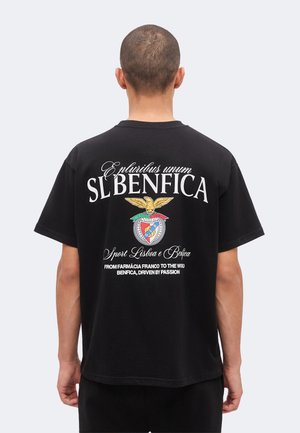 Uomo con capelli corti indossa maglietta nera SL Benfica con stemma del club e scritta bianca sulla schiena, in piedi su sfondo chiaro uniforme.