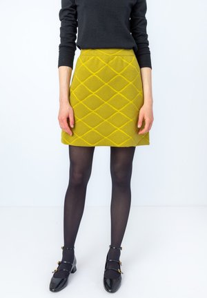 Geel gewatteerde rok met een ruitpatroon, gecombineerd met zwarte panty's en zwarte lakleren schoenen met gouden accenten.