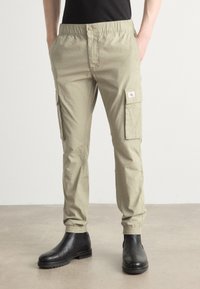 Světlé khaki cargo kalhoty s elastickým páskem, bočními kapsami a nášivkou s logem. V kombinaci s černými koženými kotníkovými botami na hladkém povrchu.