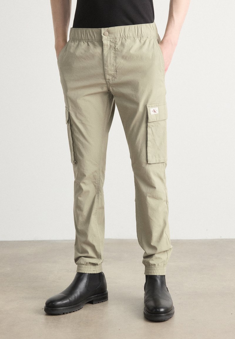 Světlé khaki cargo kalhoty s elastickým páskem, bočními kapsami a nášivkou s logem. V kombinaci s černými koženými kotníkovými botami na hladkém povrchu.