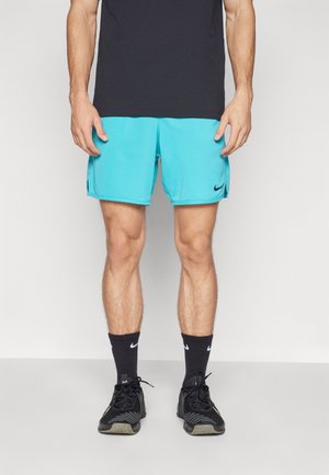 Mand iført sorte sportssko og sokker, turkise Nike-shorts og en sort T-shirt, stående mod en ensfarvet hvid baggrund.