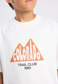 Valkoinen t-paita lyhyillä hihoilla, jossa on suuri oranssi vuoristografiikka ja rohkeat kirjaimet lukien "COLORADO TRAIL CLUB 1989" sen alapuolella.