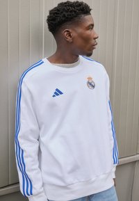 Fehér kapucnis pulóver kék Adidas csíkokkal, a mellkasán a Real Madrid logójával. Puha anyag és bordázott mandzsetta, világoskék farmernadrággal párosítva.