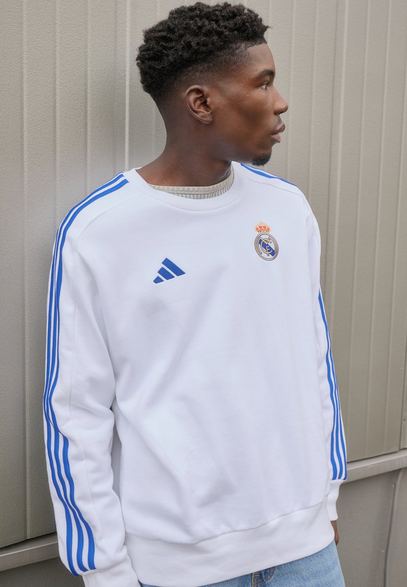 Fehér kapucnis pulóver kék Adidas csíkokkal, a mellkasán a Real Madrid logójával. Puha anyag és bordázott mandzsetta, világoskék farmernadrággal párosítva.