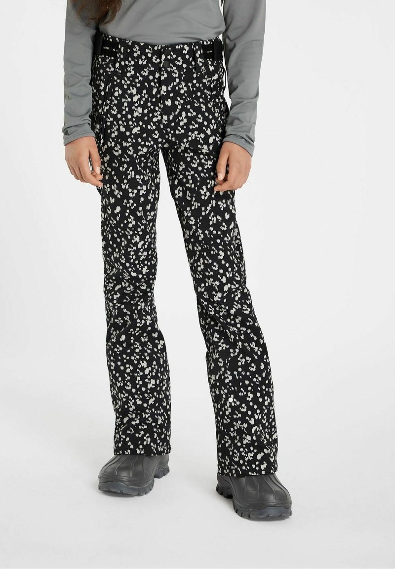 Personne portant un pantalon de ski noir avec des motifs floraux blancs, un haut à manches longues gris et des bottes de neige noires, debout sur un fond blanc.