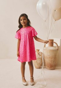 Világos rózsaszín flitteres ruha rövid, lebegő ujjakkal, ezüst lapos cipőkkel viselve. A gyermek lufikat tart, és díszedények mellett áll.