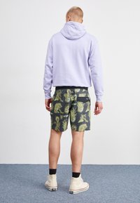 Sudadera de color púrpura claro con un bolsillo tipo canguro, combinada con pantalones cortos negros que presentan un vibrante patrón gráfico en verde y púrpura. Zapatillas blancas.