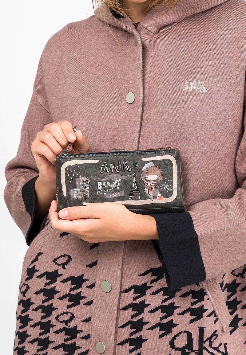 Cartera con cremallera negra que presenta ilustraciones coloridas, incluyendo la Torre Eiffel, texto y un personaje. El exterior tiene una textura suave.
