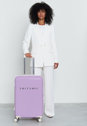 Purple hard-shell suitcase with a glossy finish, silver logo "SUITSUIT," and dual wheels. Accompanied by a model in a white suit.
