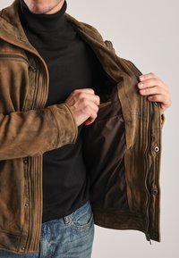 Brun læderjakke med en glat overflade og lynlåslukning. Inderlomme synlig; båret over en sort turtleneck.